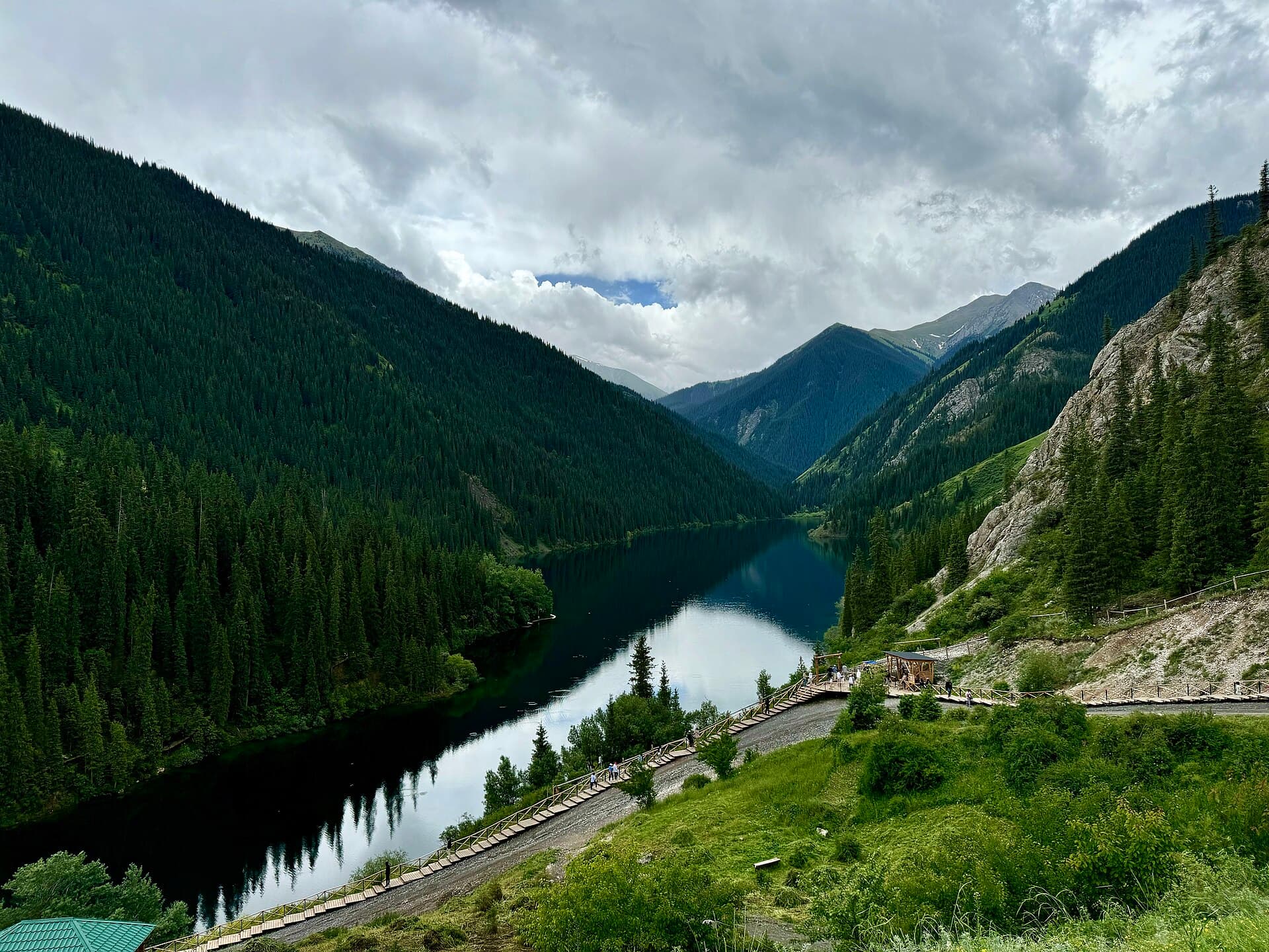 Dark spruce forest sloping to the still water of a Kolsai lake with a walkway along the shore